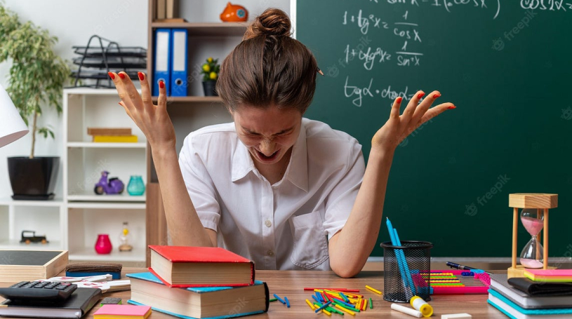 A woman is sitting at a desk in front of a blackboard with her hands in the air.