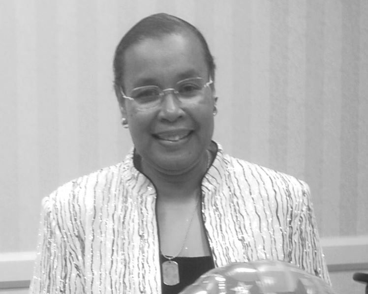 A woman wearing glasses and a striped jacket smiles in a black and white photo