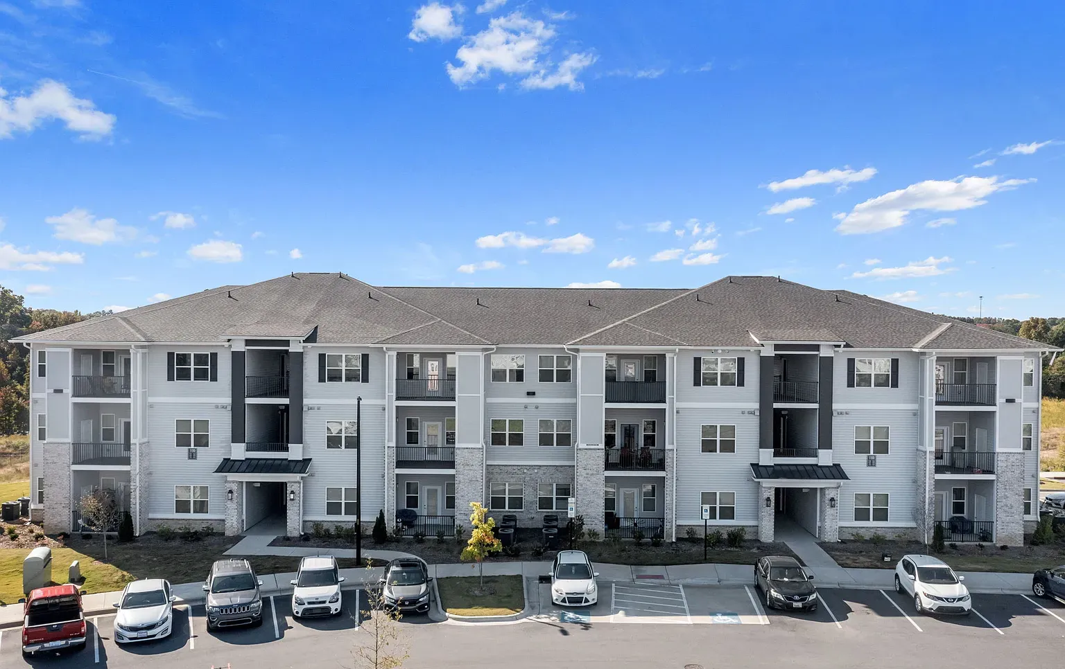 Exterior view of a modern three-story apartment building with balconies and a row of parked cars in front at Graces Reserve Apartments in Kannapolis, NC.