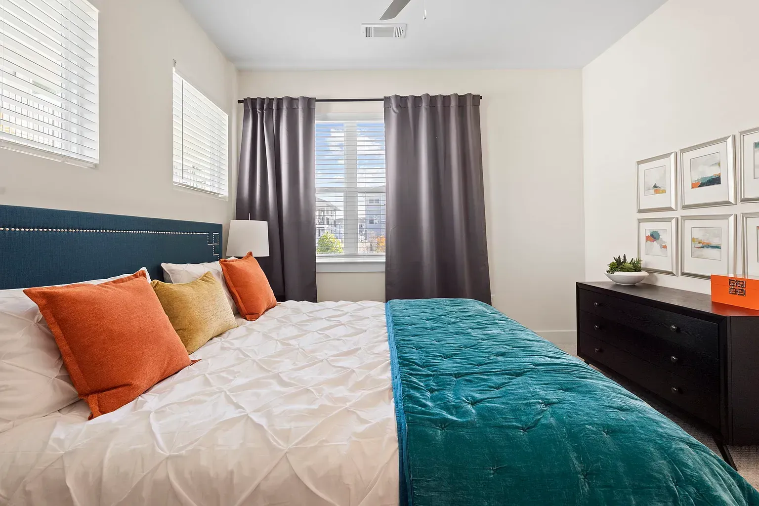 Bedroom in an apartment with a large bed, teal quilt, colorful pillows, and a dark dresser at Graces Reserve Apartments in Kannapolis, NC.