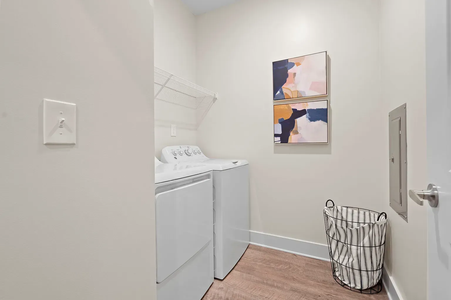 Laundry room with a washer and dryer, wire shelf, and a striped laundry basket at Graces Reserve Apartments in Kannapolis, NC.