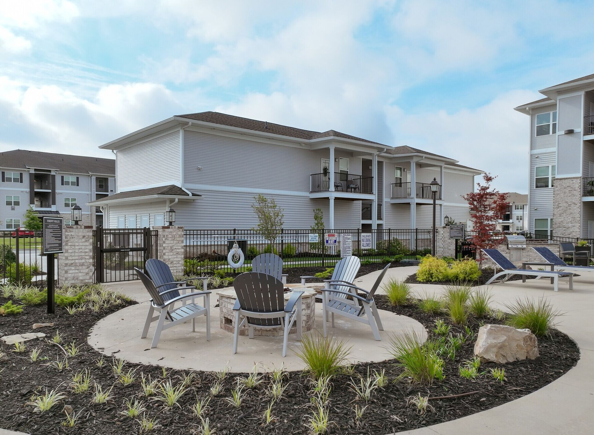 Outdoor seating area with chairs around a fire pit in front of apartment buildings under a blue sky.