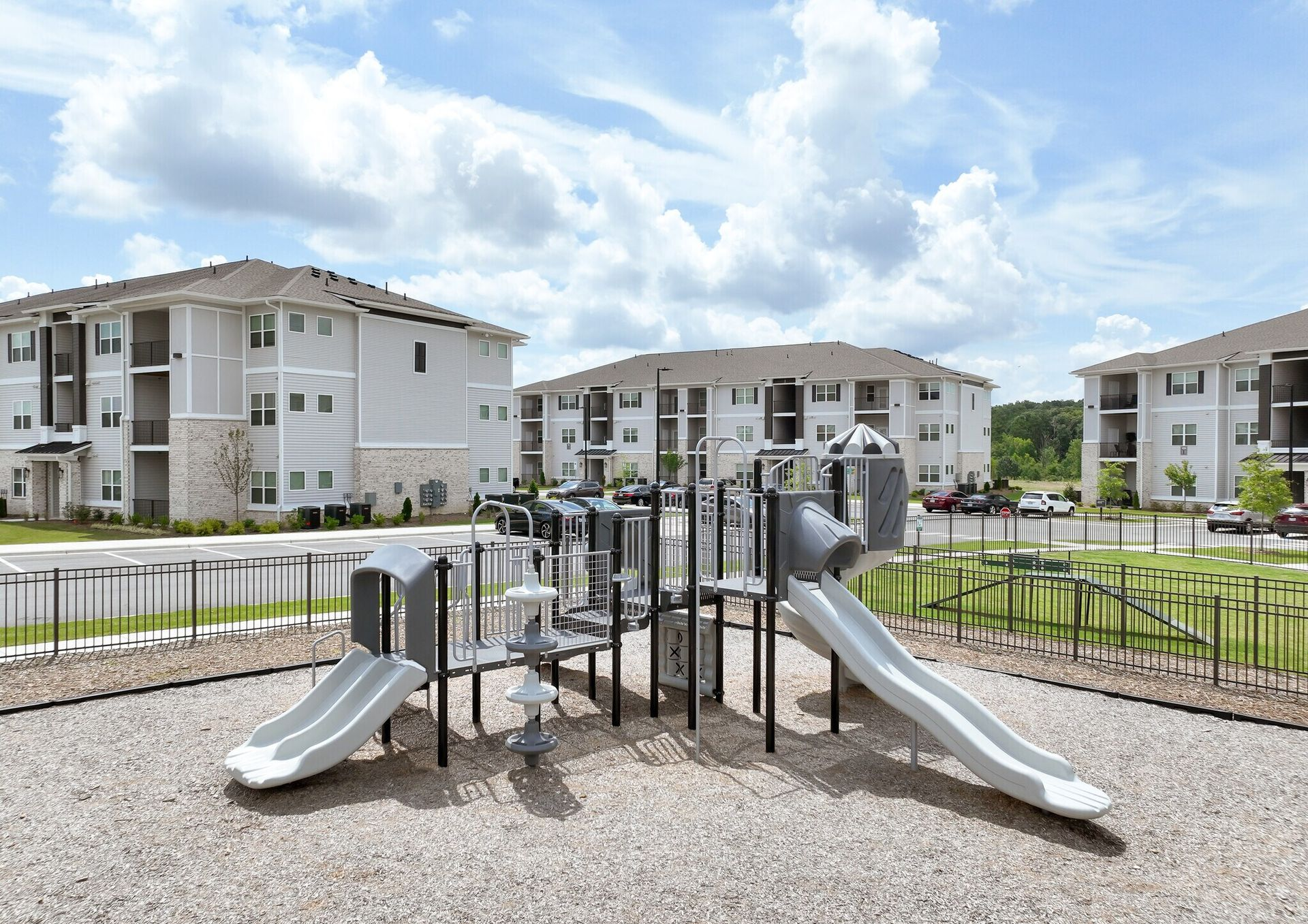 Playground with gray slides and climbing structures in front of apartment buildings under a cloudy sky.