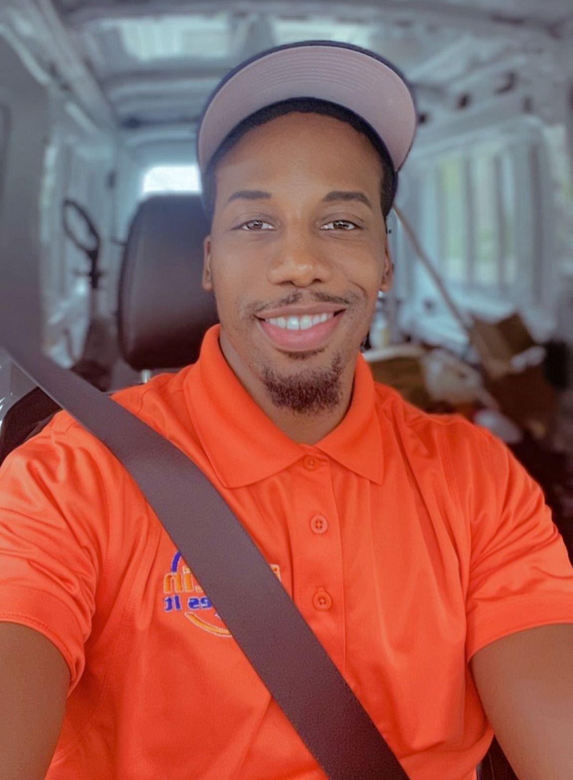 A man wearing an orange shirt and a hat is sitting in a van.