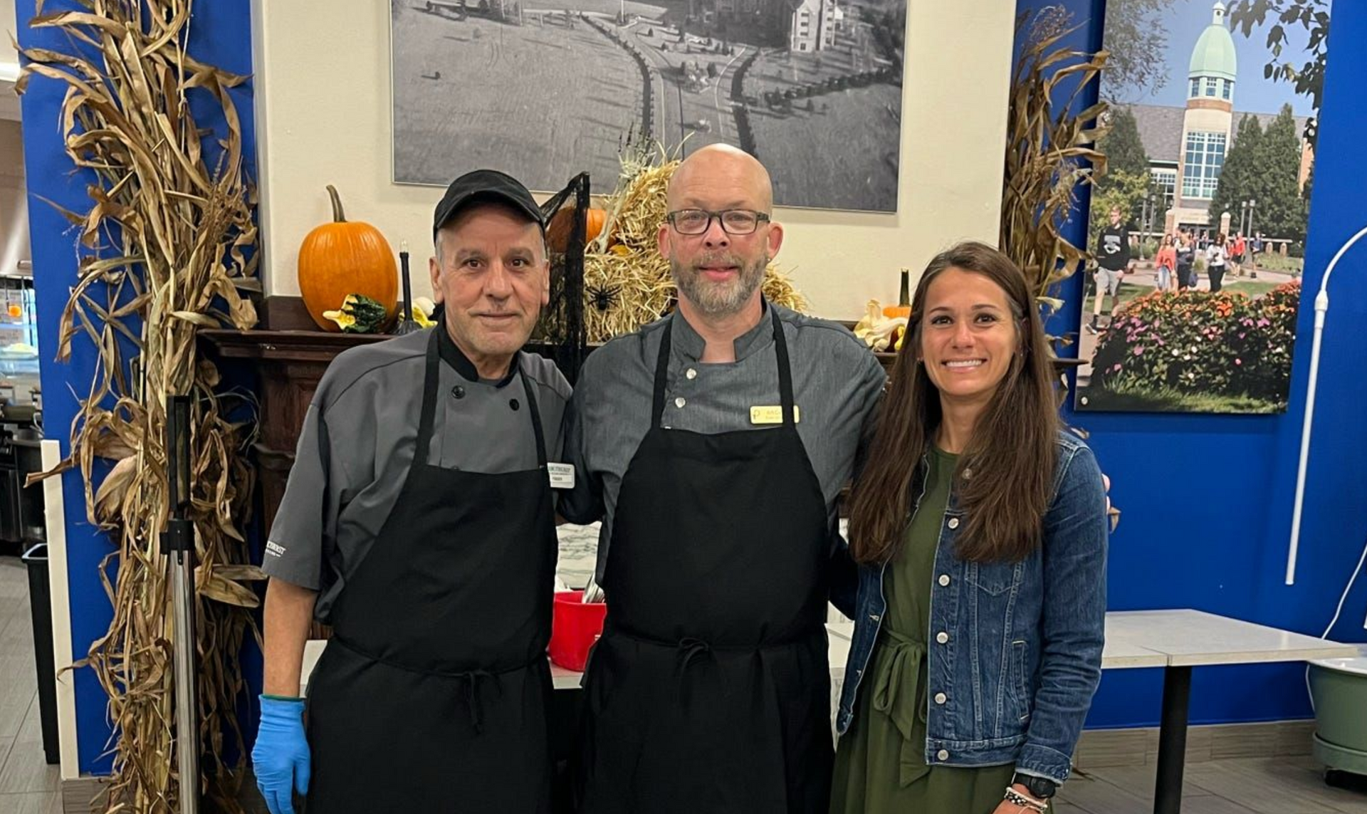 Two individuals in chef uniforms and a woman stand together in a decorated room with pumpkins and wall art.