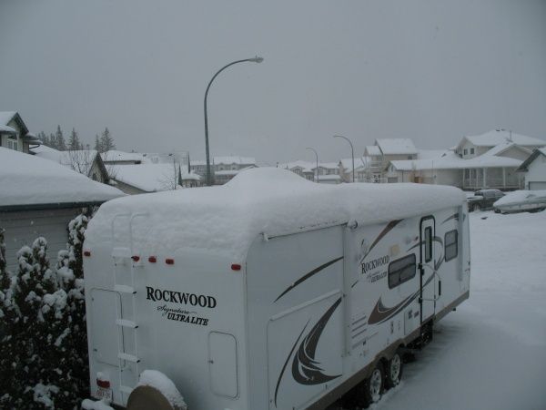 Snow-covered RV parked in a residential area; rooftops and trees also covered in snow; cloudy, overcast sky.