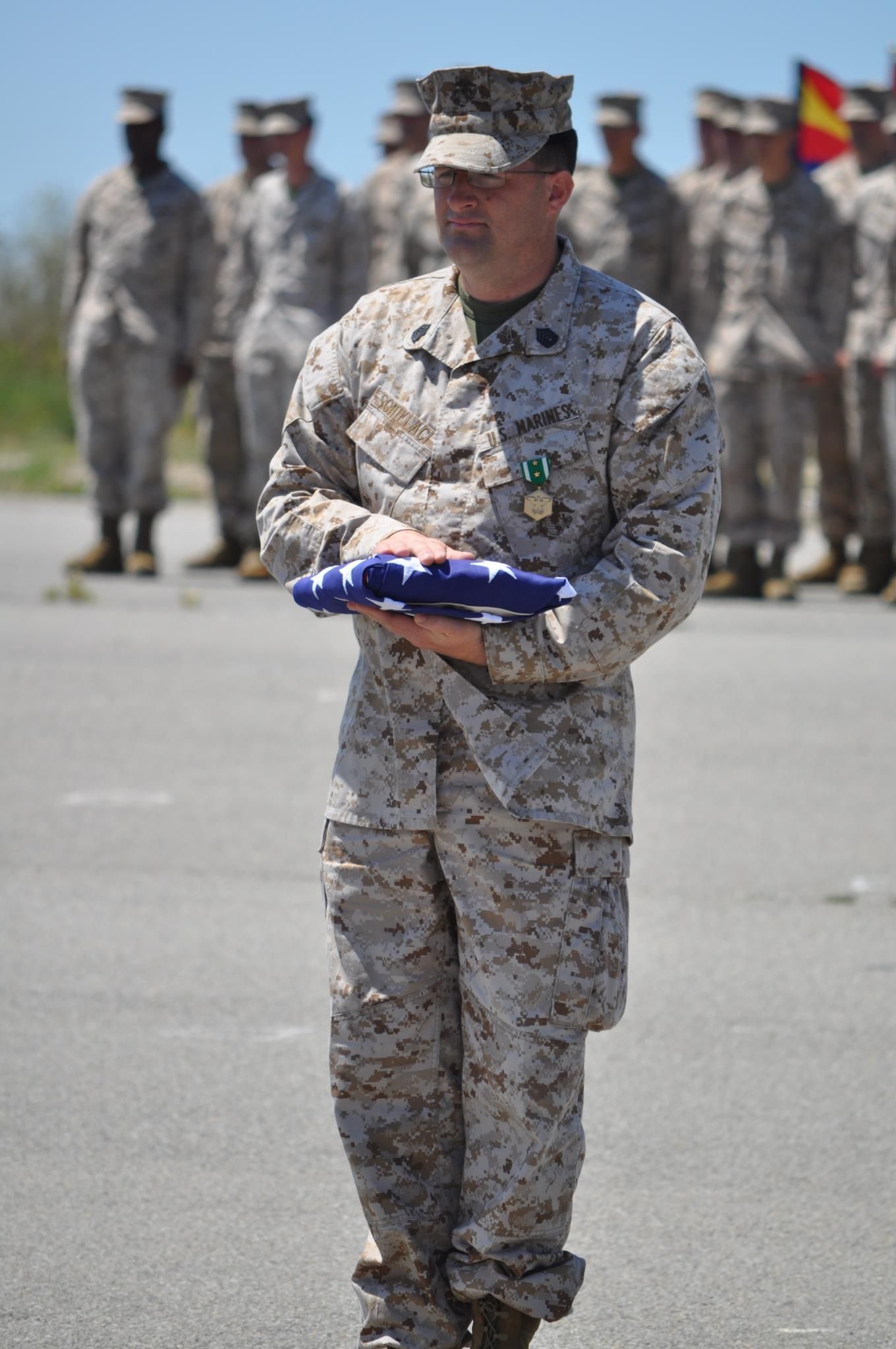 Marine holding a folded American flag during a ceremony, other Marines in the background.