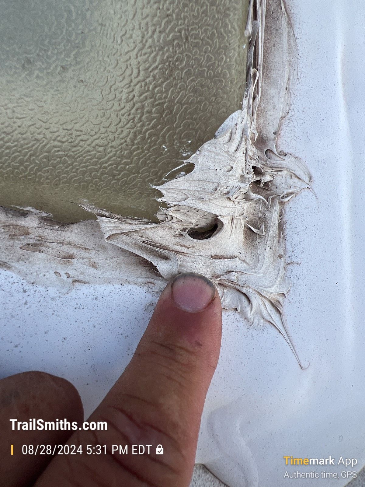 A person's finger pointing at peeling sealant around the edge of a window, revealing wear and tear on a white surface.
