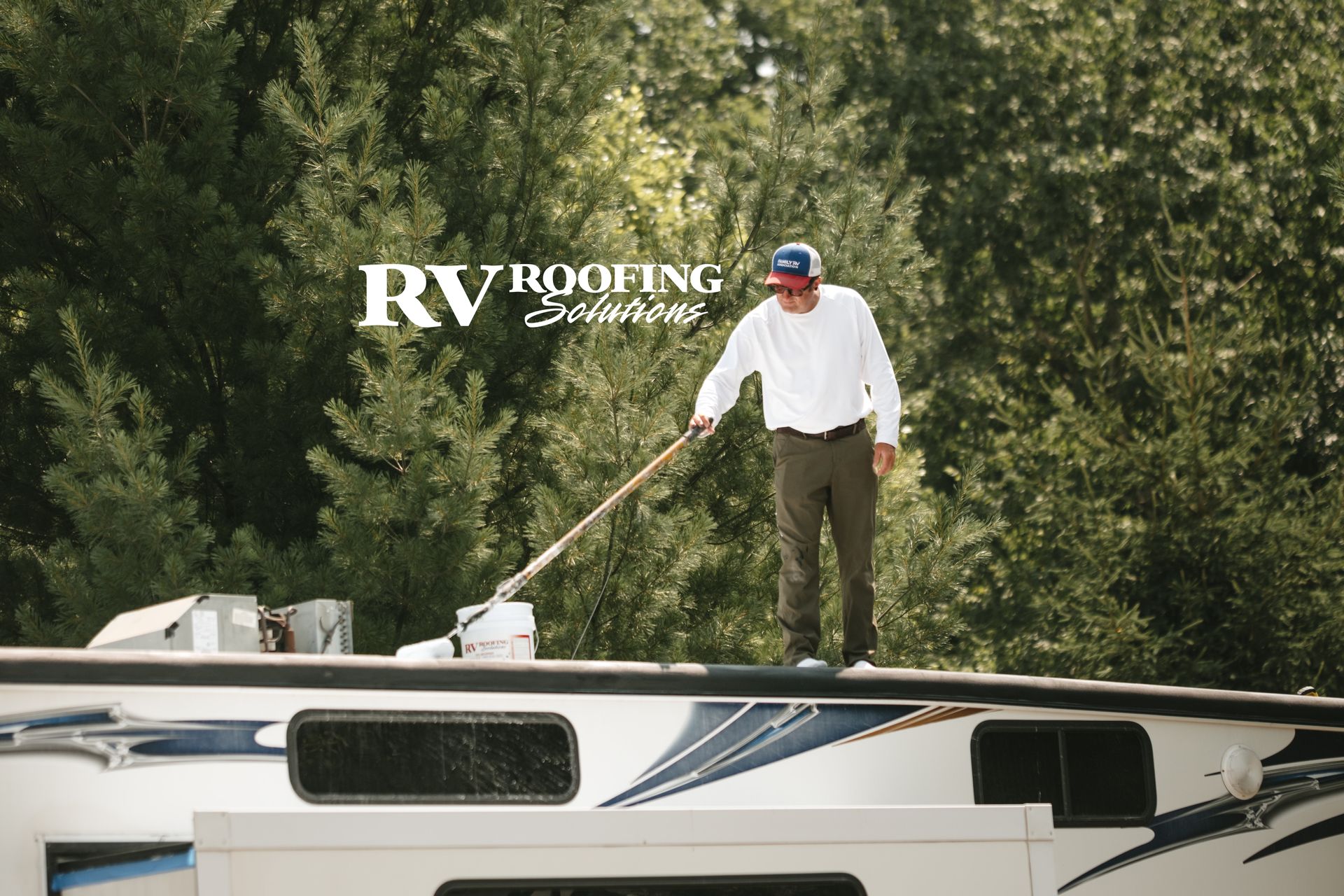 Man in white shirt paints an RV roof, with green trees in the background.