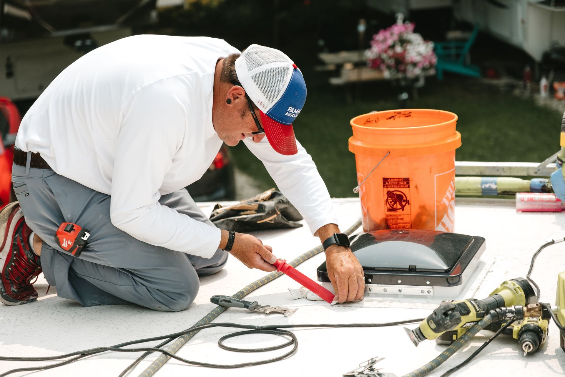 Man on RV roof, wearing hat, working with tools near orange bucket.
