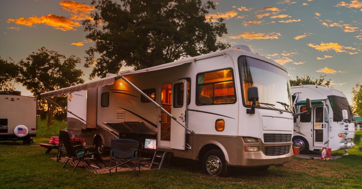 A white RV in an RV park. The RV's awning is deployed and covering several lawn chairs and picnic blankets.