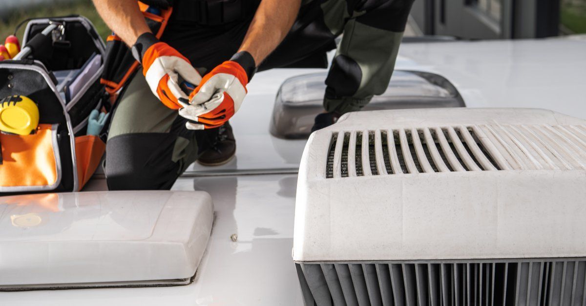 A technician in black and orange clothes works on the air conditioning unit that is on top of an RV's roof.