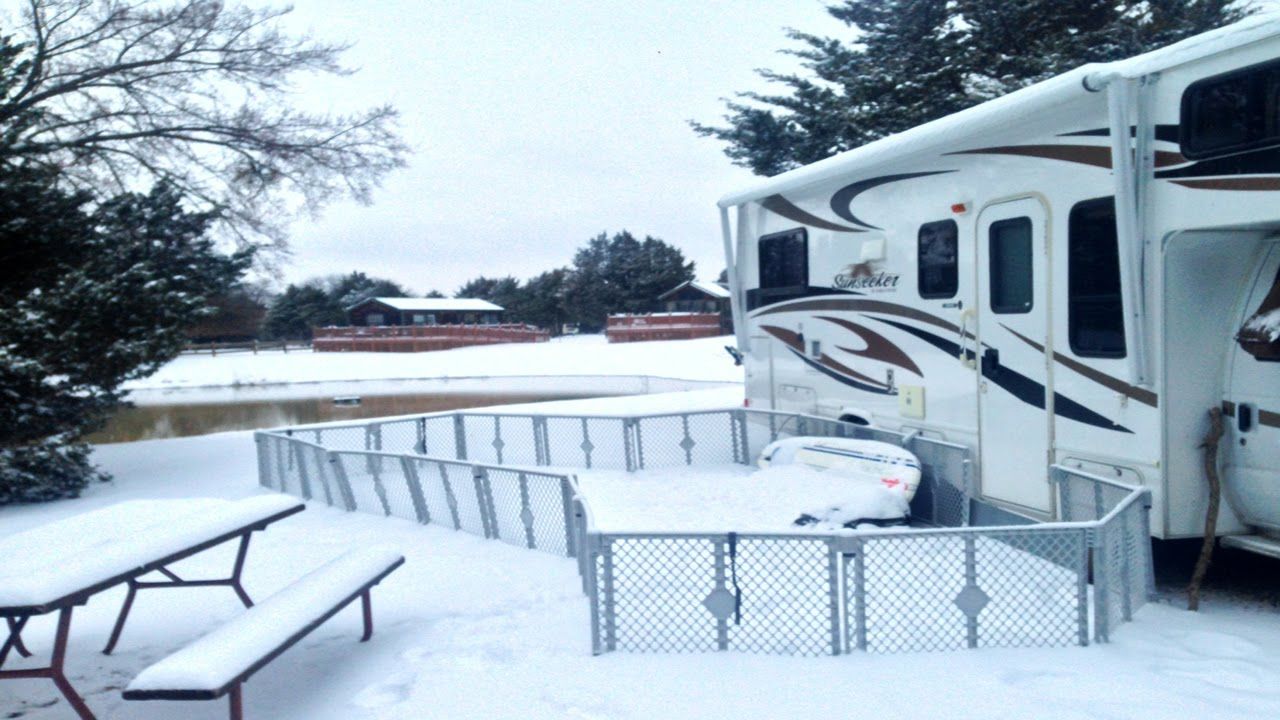 Snowy RV campsite with RV, picnic table, and fence. Overcast day, snow covers everything.
