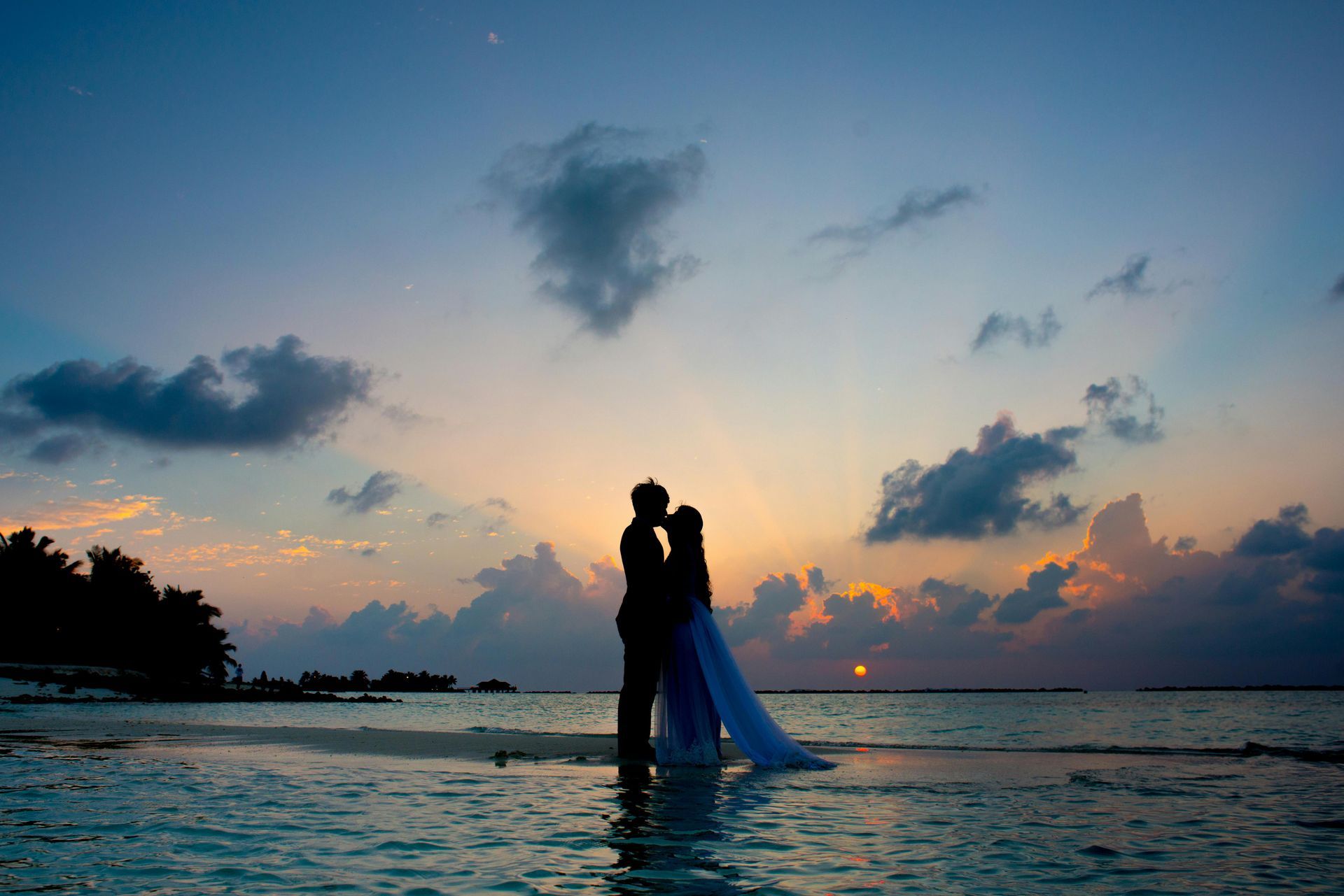 A bride and groom are kissing on the beach at sunset.