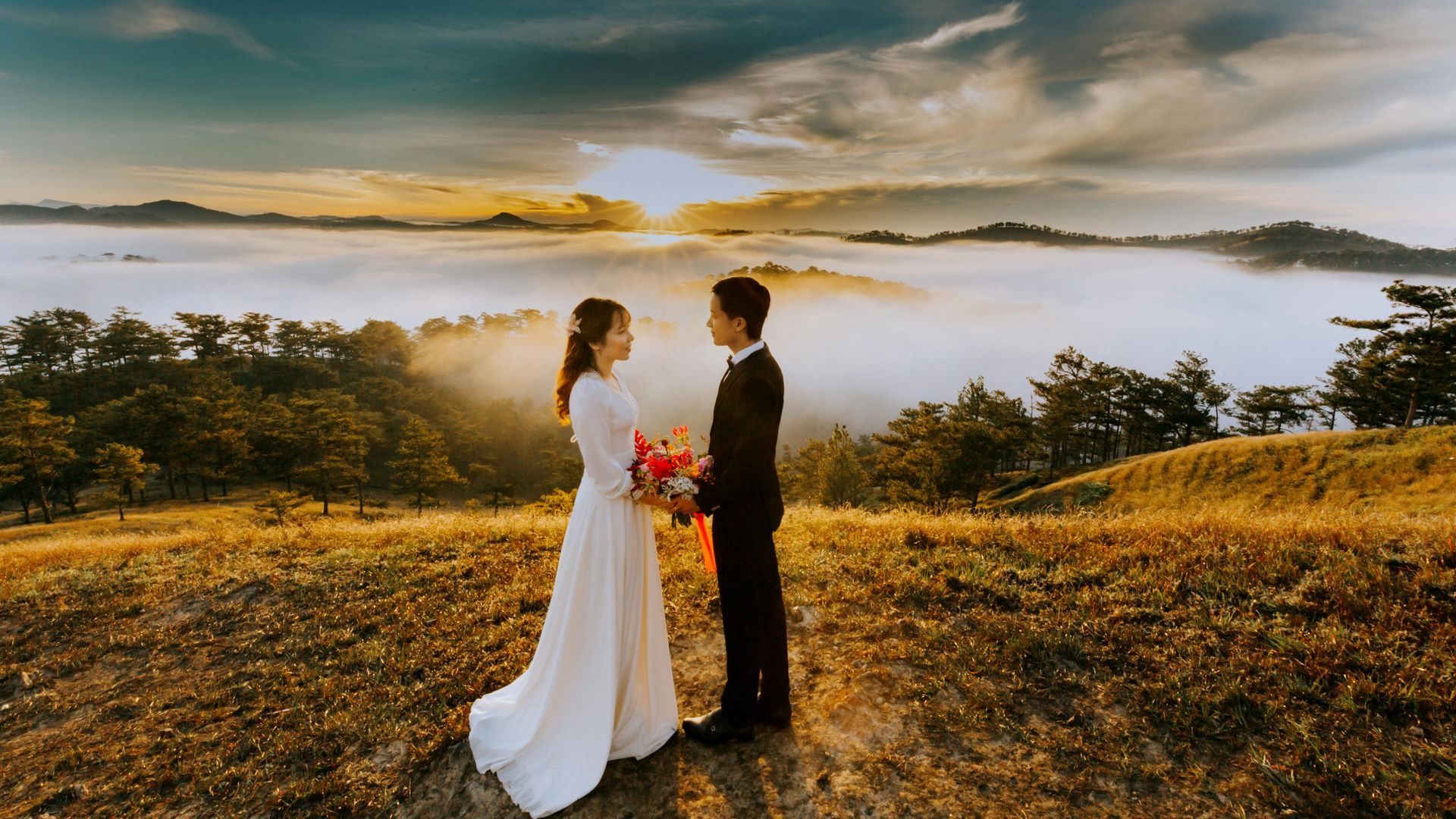A bride and groom are standing on top of a hill holding hands.