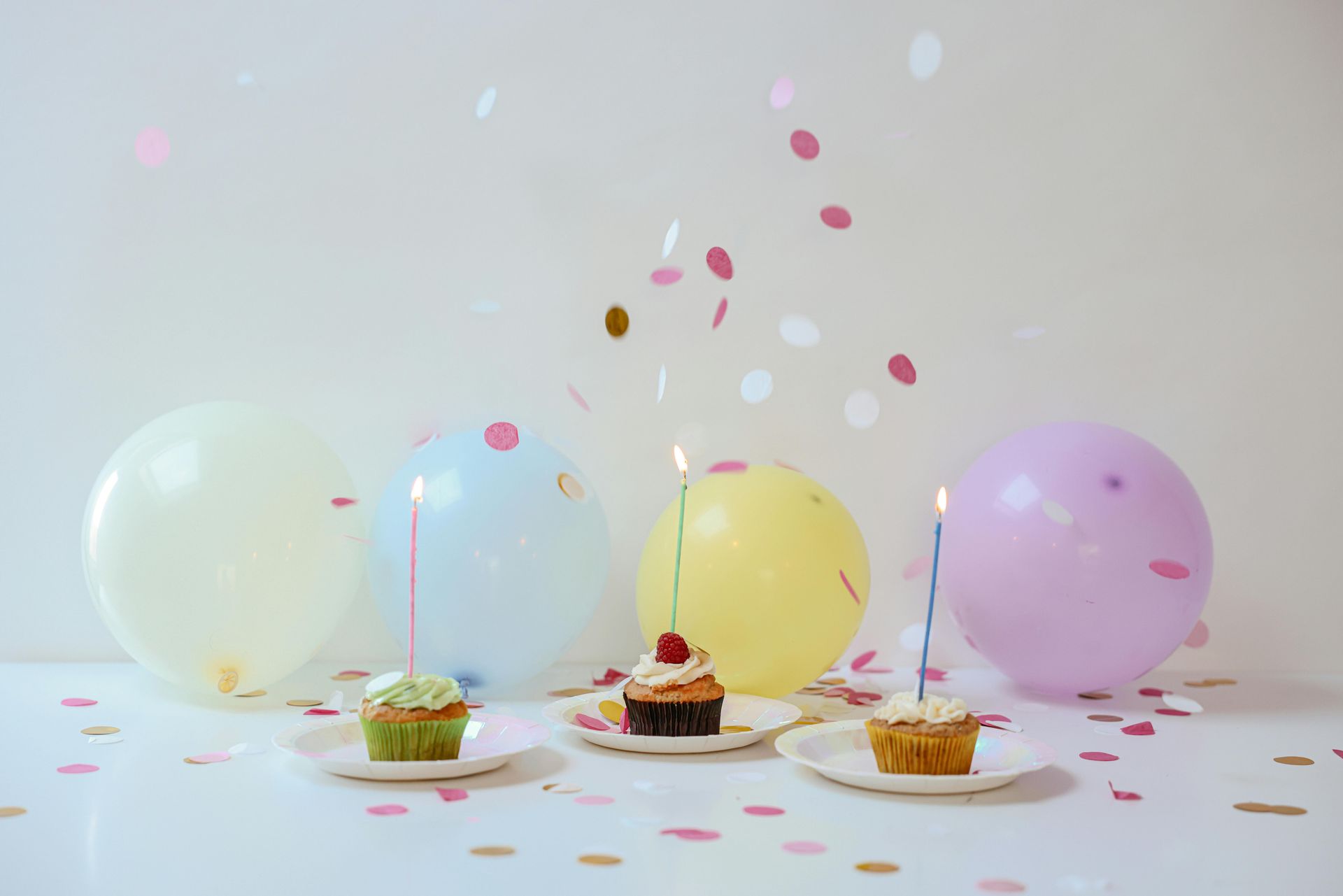 Three cupcakes with candles on them are sitting on plates next to balloons and confetti.
