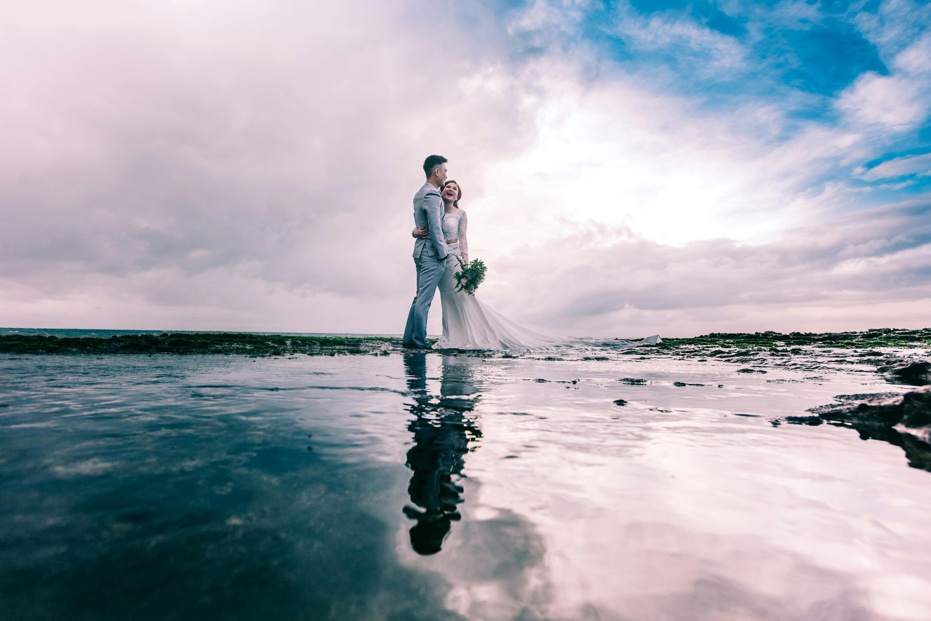 A bride and groom are standing in the water on a beach.