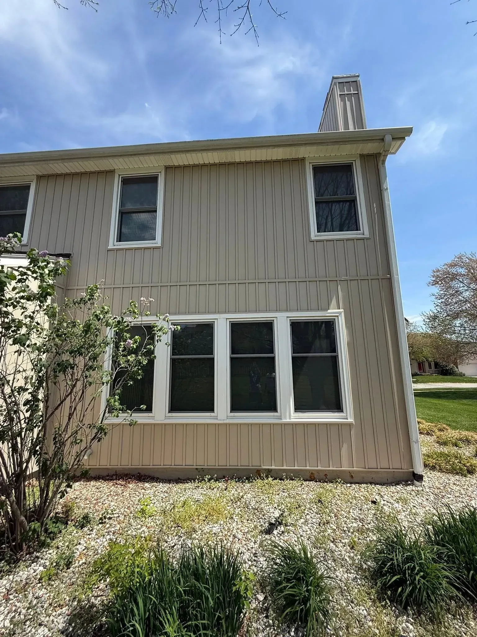 Tan two-story house exterior with vertical siding, several windows, and a chimney on a sunny day.