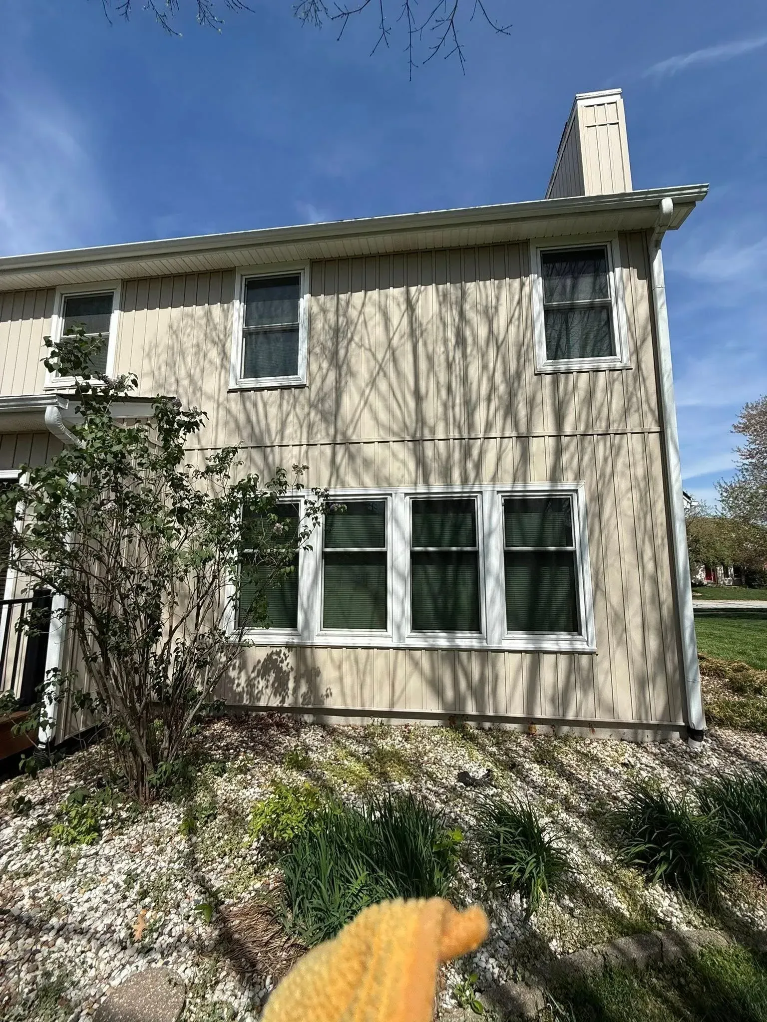 Beige two-story house with brown window shutters, a chimney, and a garden in the foreground on a sunny day.
