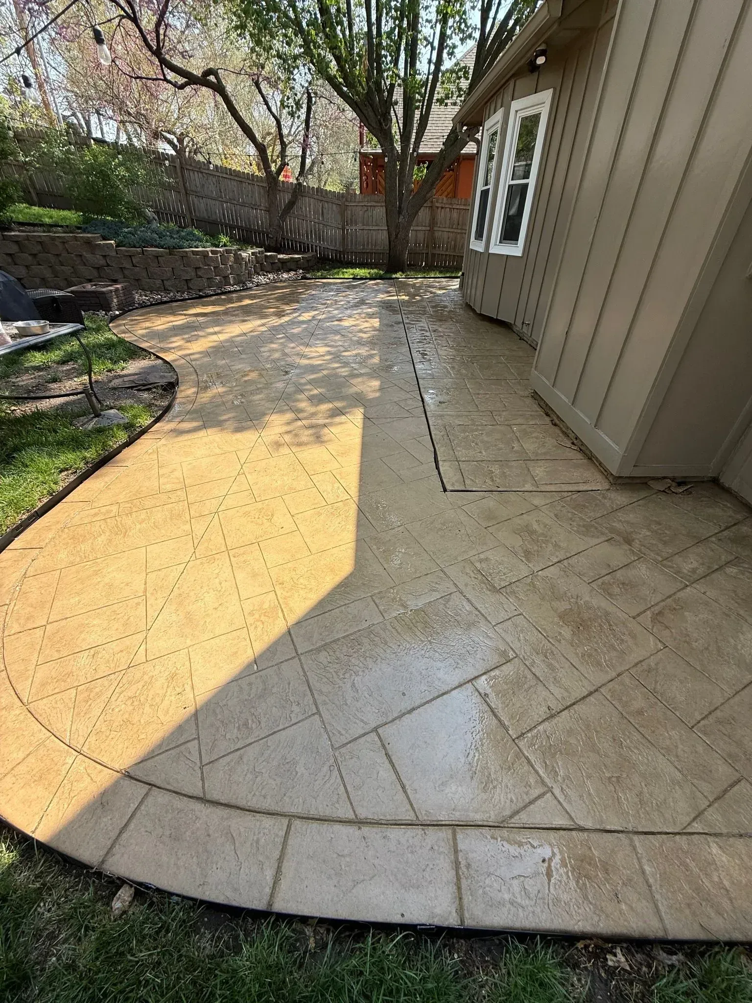 A paved stone patio with a dark border runs alongside the beige exterior wall of a house, set in a sunny backyard.