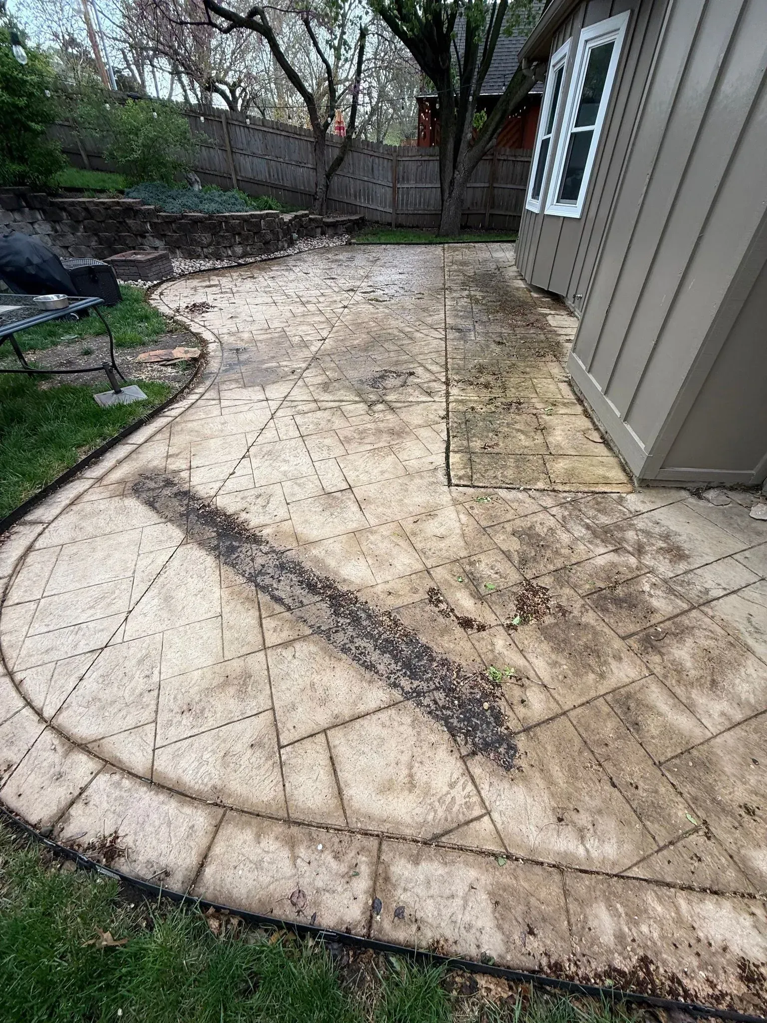 A dirty, patterned concrete patio in a backyard, showing visible stains, debris, and wear next to a house siding.
