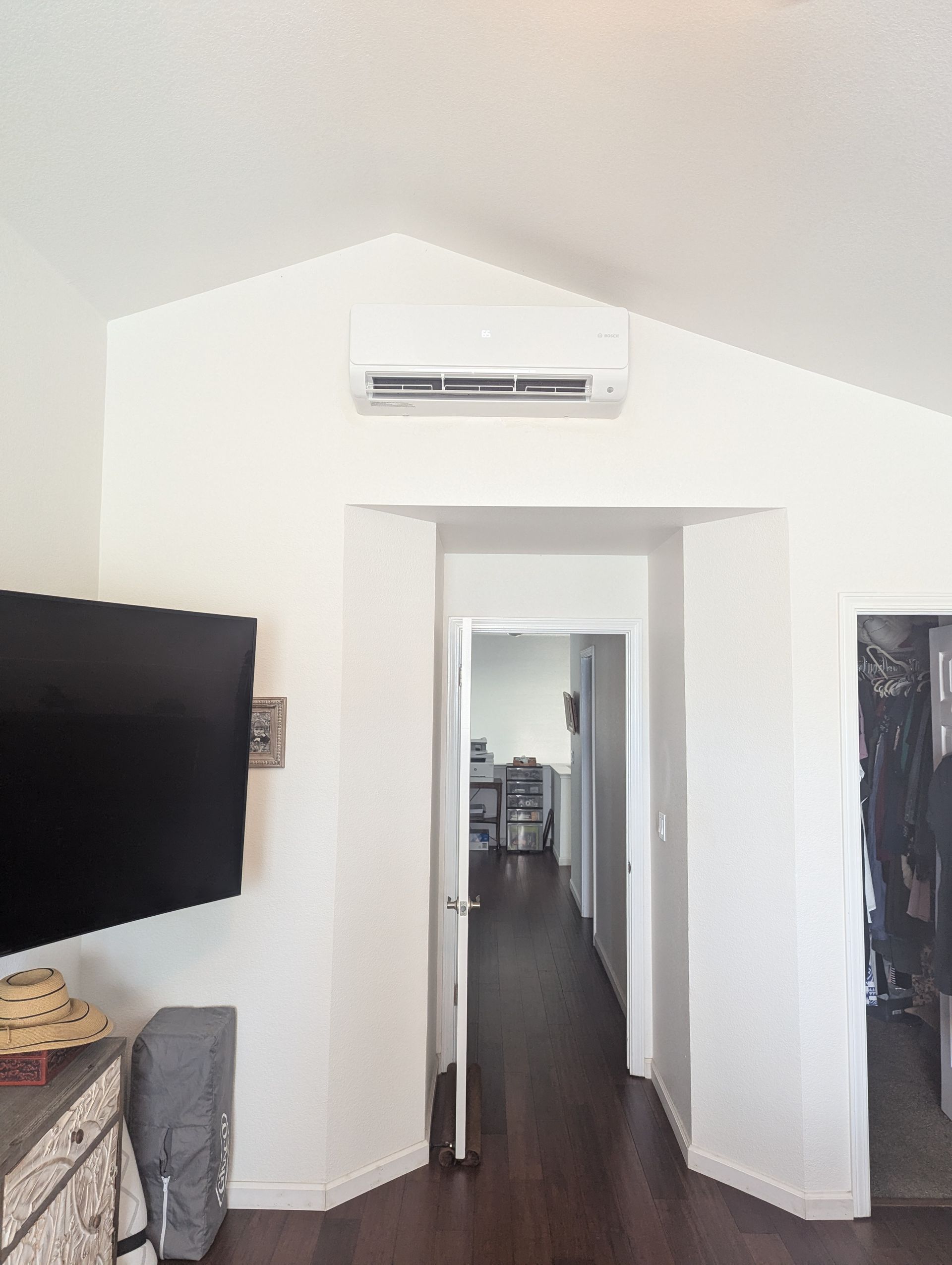 A white interior wall with a mounted air conditioner, a doorway, and a television. Dark wood floors and a bright hallway are visible.