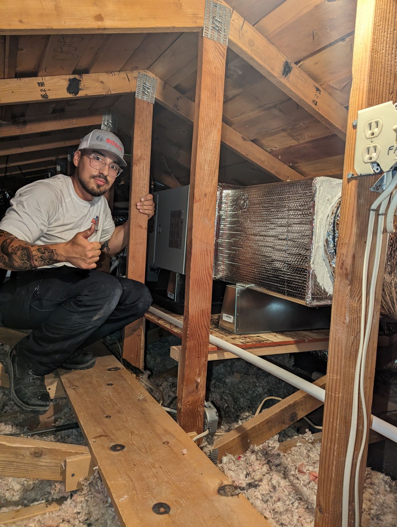 Man in an attic gives a thumbs-up next to HVAC unit. He is wearing a cap and has arm tattoos. Attic is dusty with exposed wooden beams.