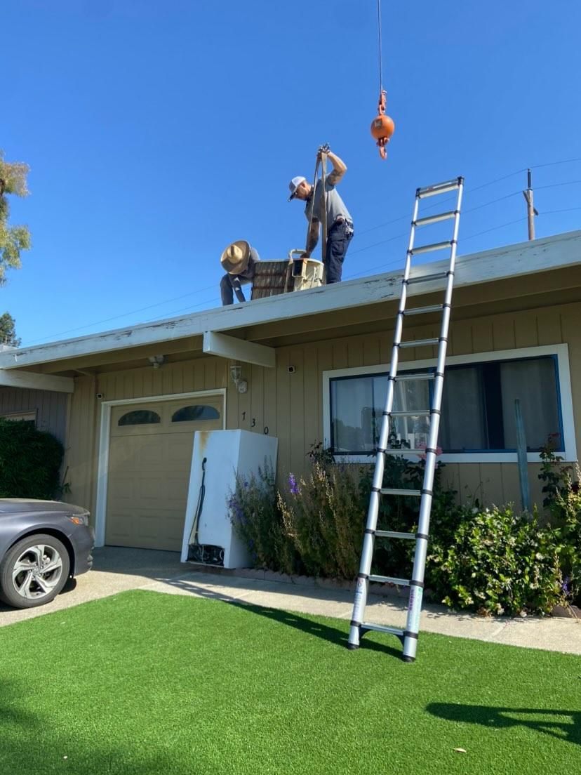 Two workers on a roof near a ladder and a lifted object. A refrigerator sits on the lawn. Sunny day.