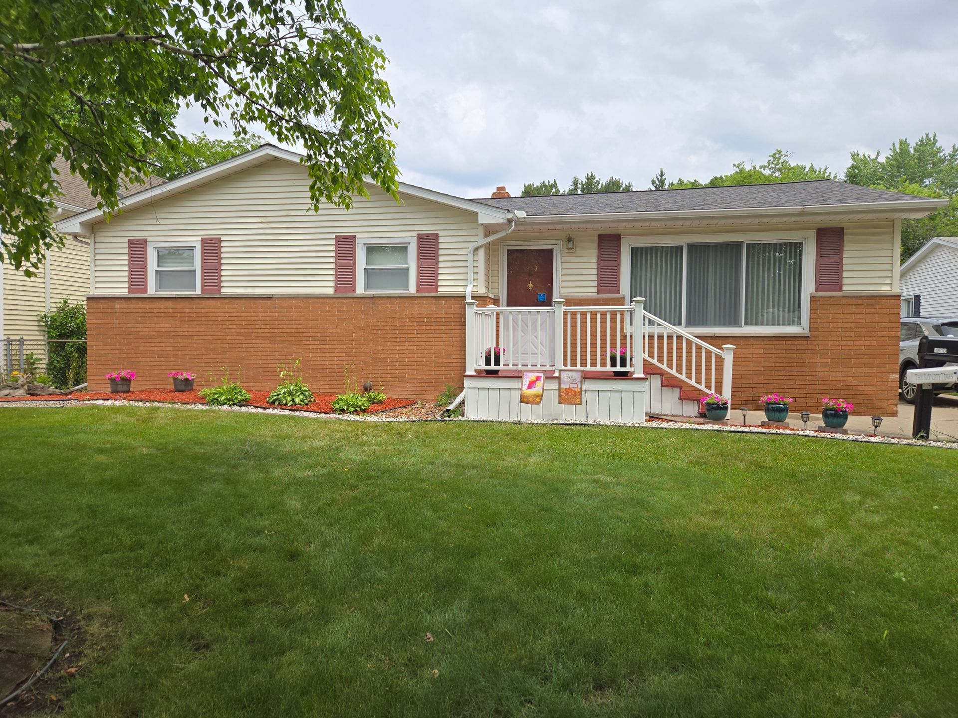 Ranch-style house with tan siding, red shutters, and brick facade; small front porch with white railing, set on green lawn.