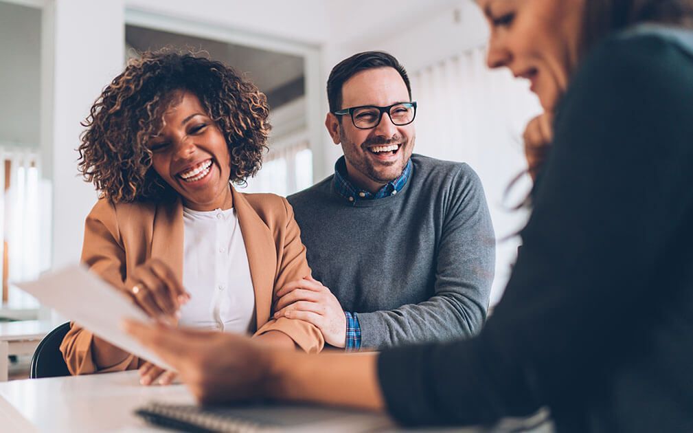 Couple smiles as they review documents with a financial advisor. Indoors, neutral tones.