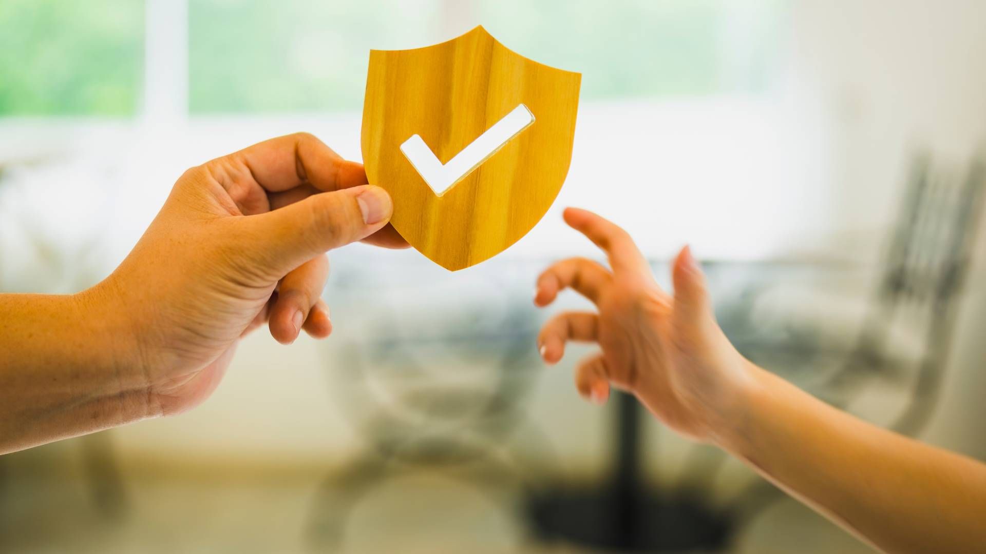 Hands holding a golden shield with a white checkmark, reaching towards another hand.