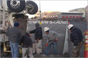 Workers pouring concrete from a truck into a form in a paved area, with a large building in the background.