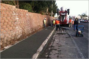 Construction workers pouring concrete for a sidewalk next to a retaining wall.