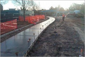 Newly poured concrete walkway under construction; orange safety fencing on the side, worker in the distance.