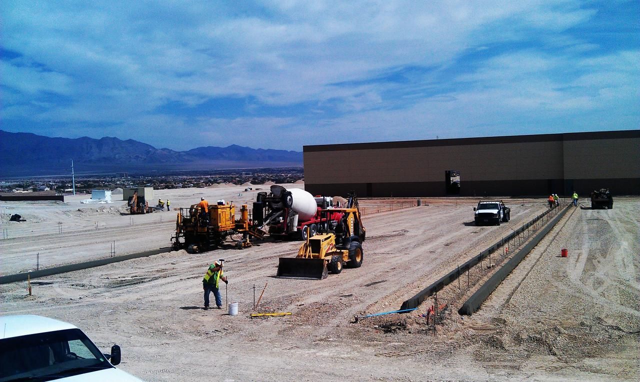 Construction site with machinery and workers pouring concrete curbs under a cloudy sky.