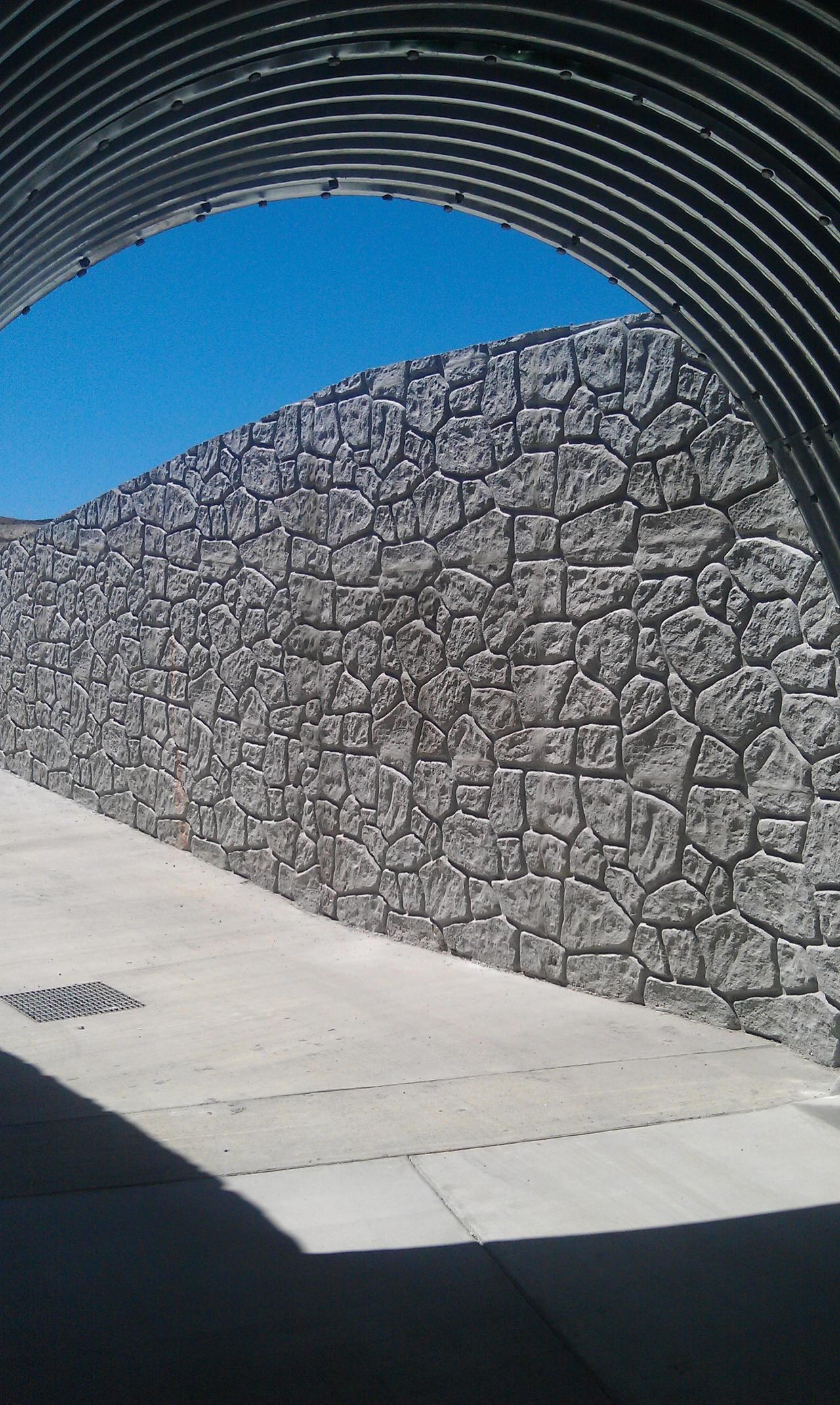 Stone wall and sidewalk framed by corrugated metal archway, blue sky visible.