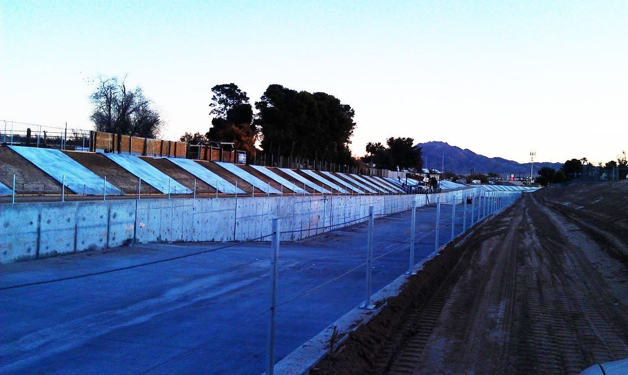 Concrete drainage channel with sloped sides, fence, and dirt path under a twilight sky.