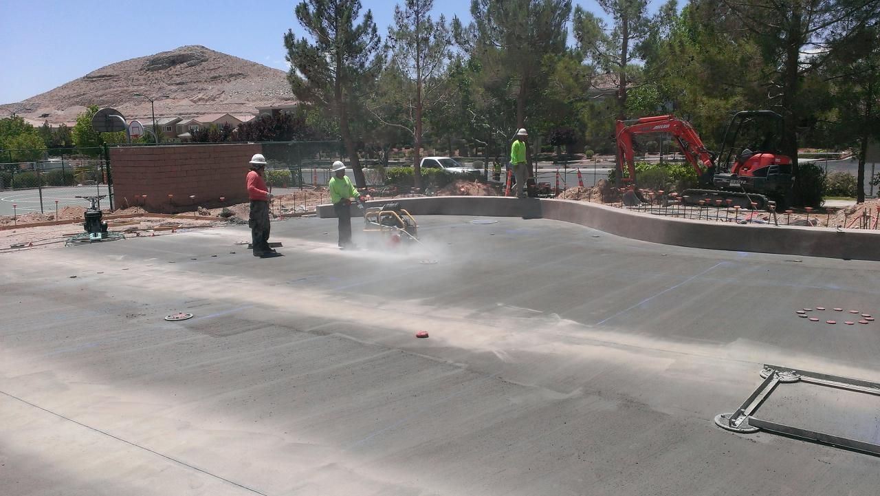 Construction workers grinding concrete in outdoor setting. Trees, rock mountain, and small excavator visible.