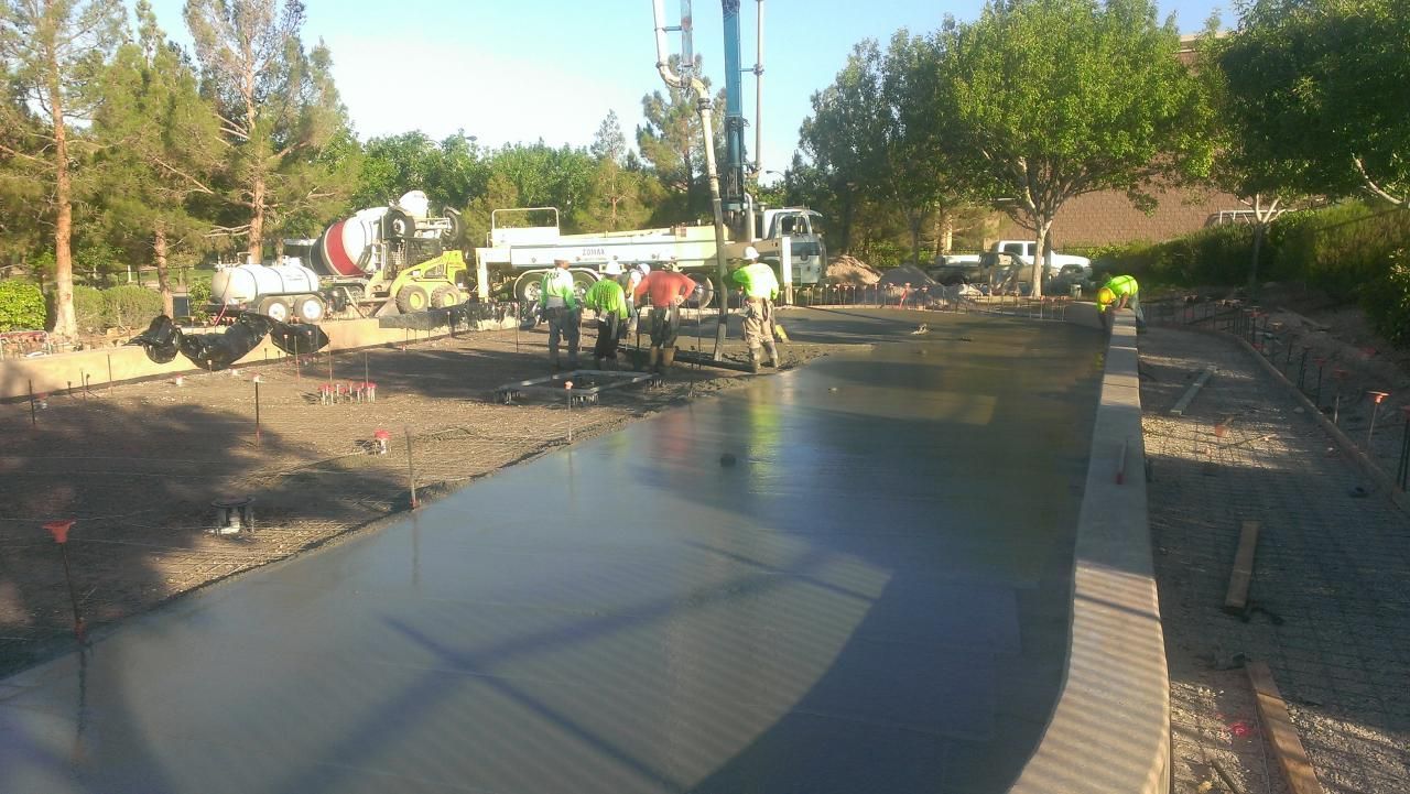 Workers pouring concrete, construction site, daytime. Cement truck and pump, flat grey surface, green safety vests.