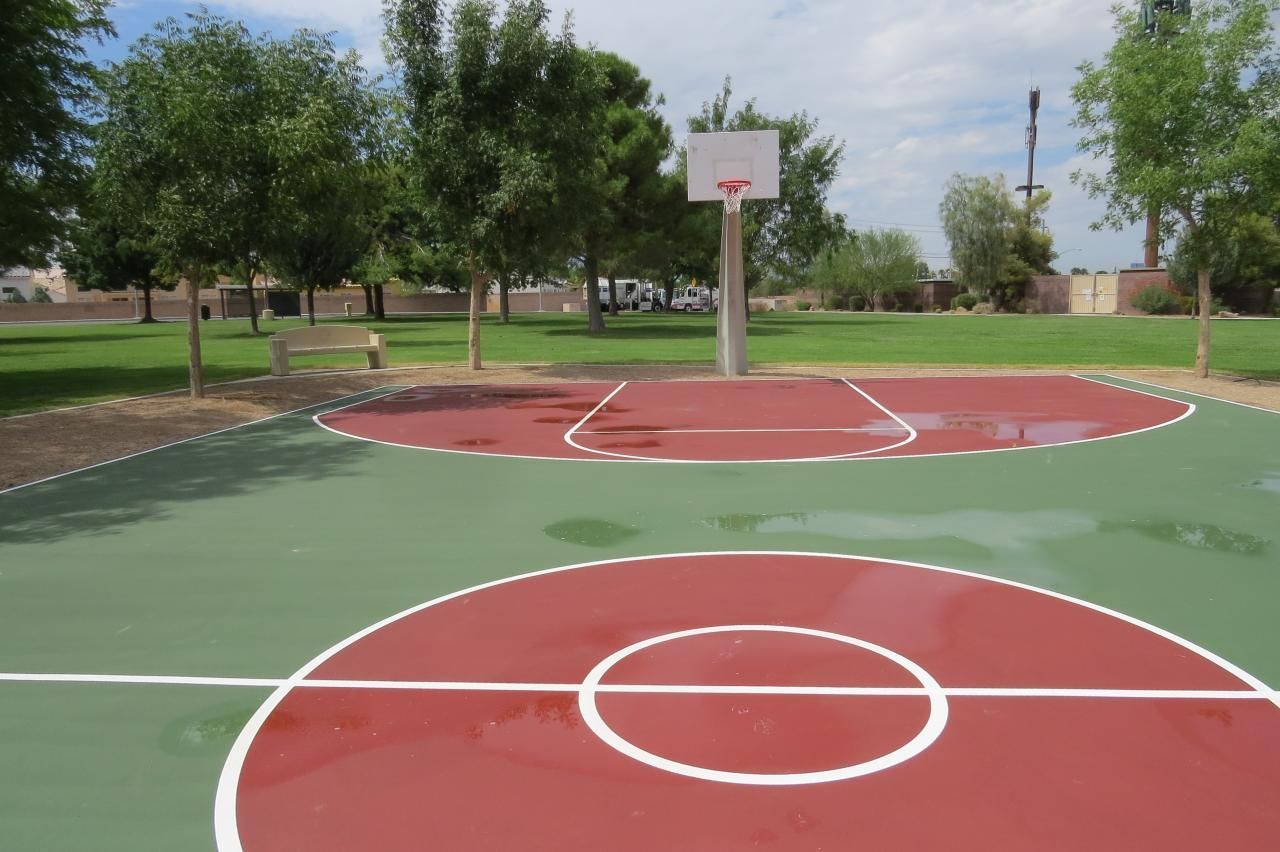 Outdoor basketball court, green and red painted surface, hoop with white backboard, trees in background.