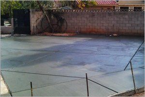 Newly poured concrete slab in a yard, bordered by a wall and gate, metal rebar stakes.