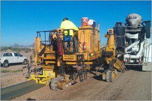 Yellow machinery laying a concrete pipeline, workers present, with a cement truck in the background, outdoors on a sunny day.