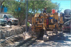 Construction workers operating a yellow machine laying concrete on a street. A concrete truck and vehicles are nearby.