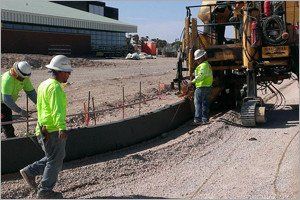 Construction workers in safety vests installing a concrete curb near a building, using machinery.