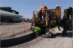 Construction workers operating a yellow machine building a concrete curb on a dirt road.