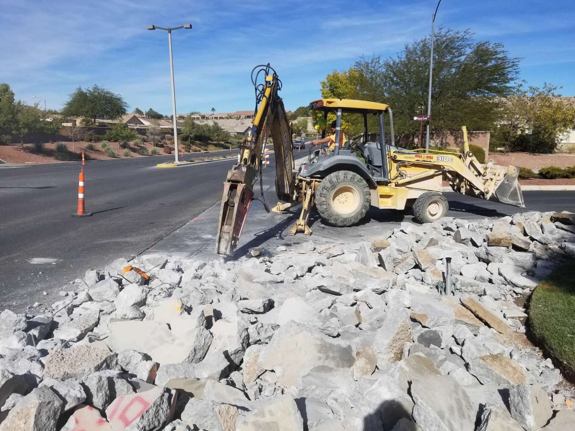 A yellow backhoe breaking up concrete on a road, with debris scattered.