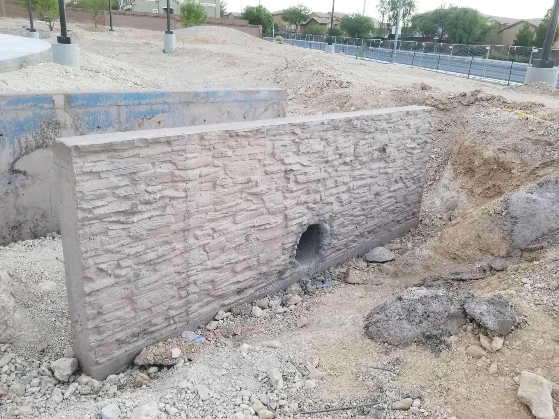 Concrete retaining wall with a hole, surrounded by dirt and gravel.