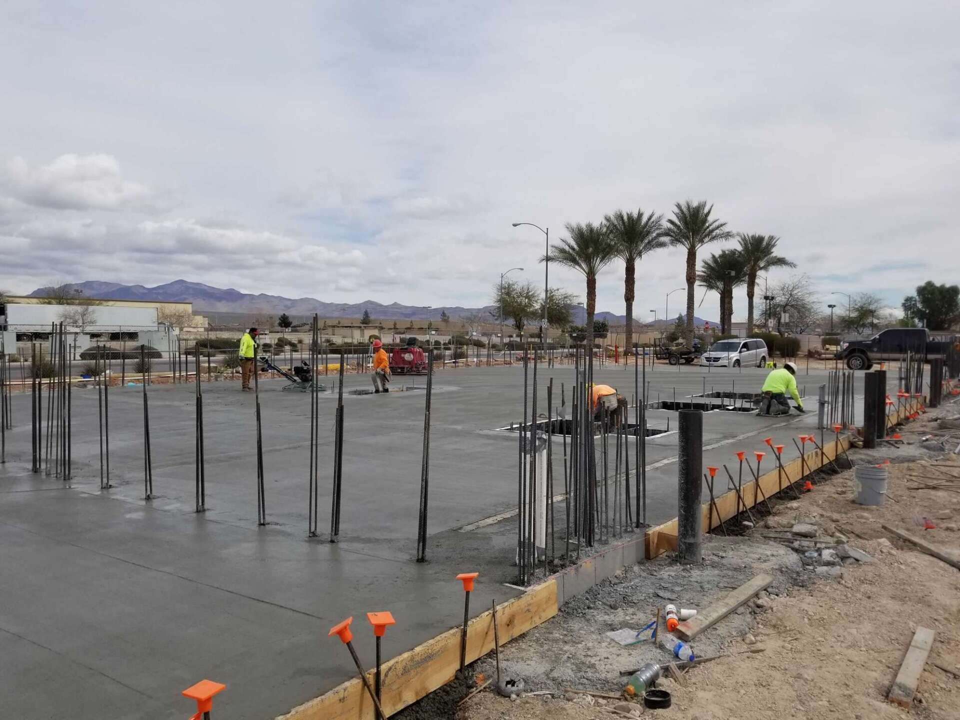 Construction site with workers pouring concrete. Rebar stands upright. Palm trees and cloudy sky in the background.