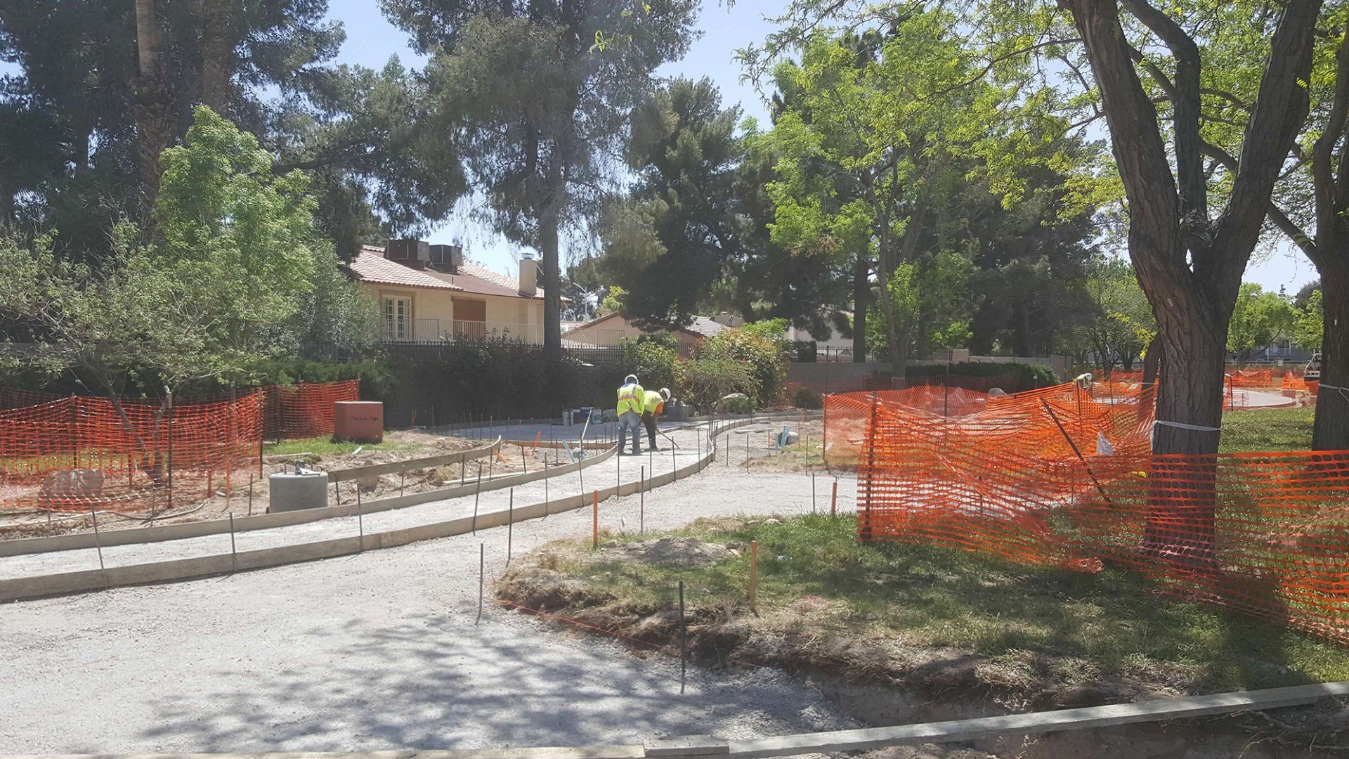 Construction site with orange safety fencing and workers near a curved pathway in a park.