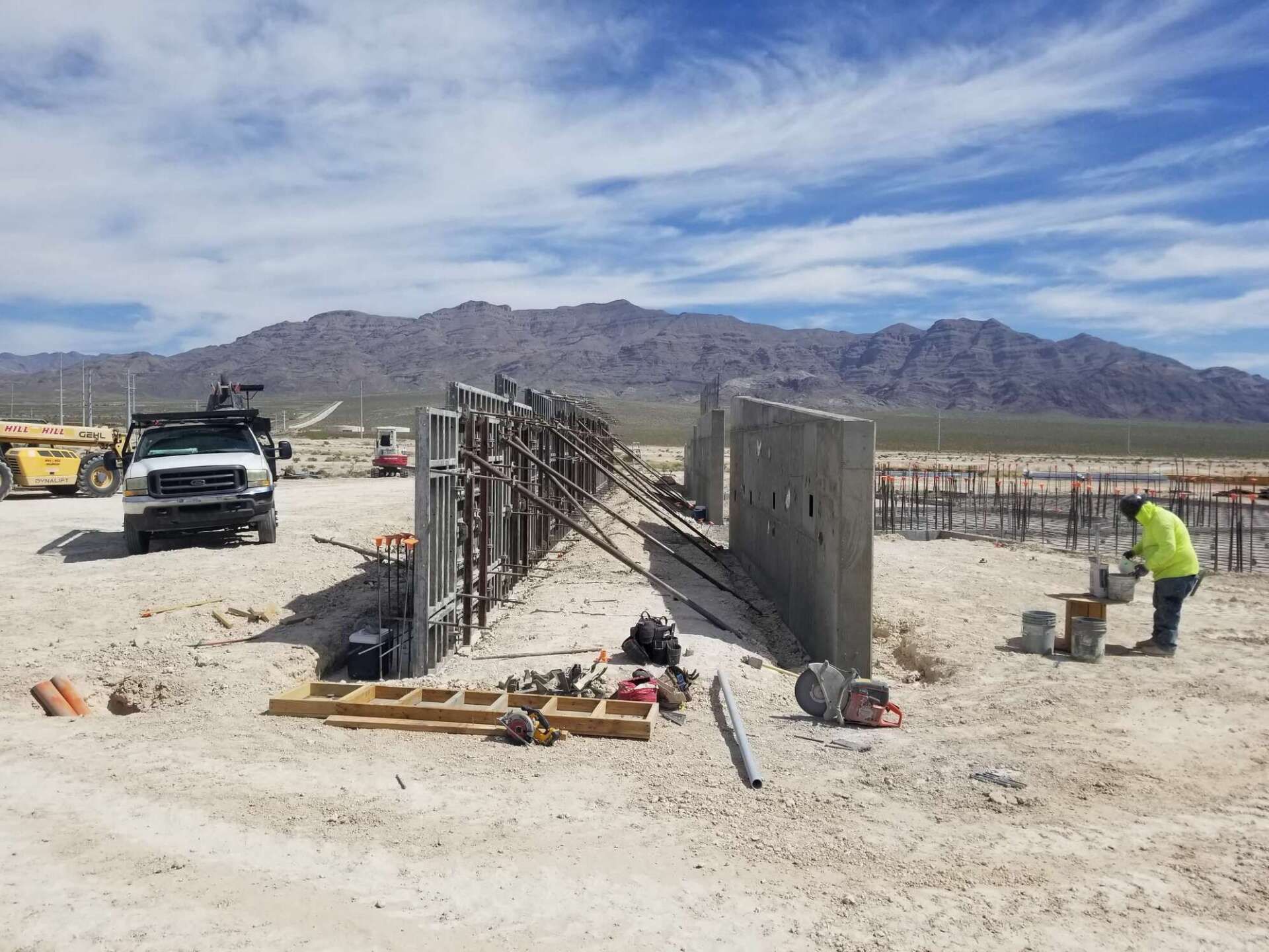 Construction site: wall formwork and worker pouring concrete, mountains in the background, sunny day.